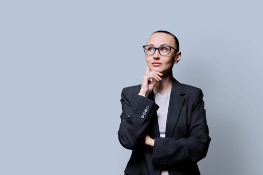 Portrait of thinking serious 30s business woman on grey studio background. Confident female in glasses suit looking to side at space for image text copy space. Business work teaching job career people