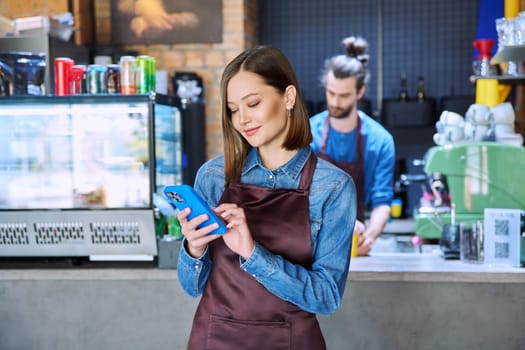 Confident successful young woman service worker in apron holding smartphone in restaurant cafeteria coffee shop pastry shop. Small business, staff, occupation, entrepreneur owner, work concept