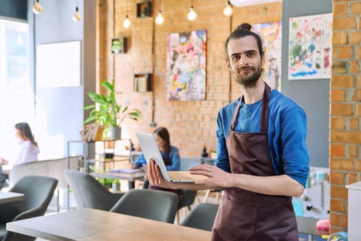 Successful young man service worker owner in apron holding using laptop computer looking at camera in restaurant cafeteria coffee pastry shop interior. Small business staff occupation entrepreneur work