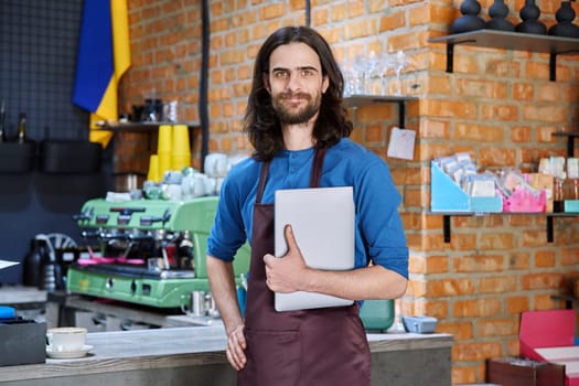 Young man in apron, food service worker, small business owner entrepreneur holding laptop looking at camera near counter of coffee shop cafe cafeteria. Staff, occupation, successful business, work