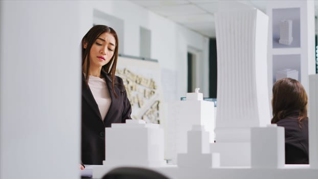 At design table, engineer reviews schematics and 3D construction plans in order to develop manufacturing layout for urban housing project. Woman evaluates sizes and components. Handheld shot.