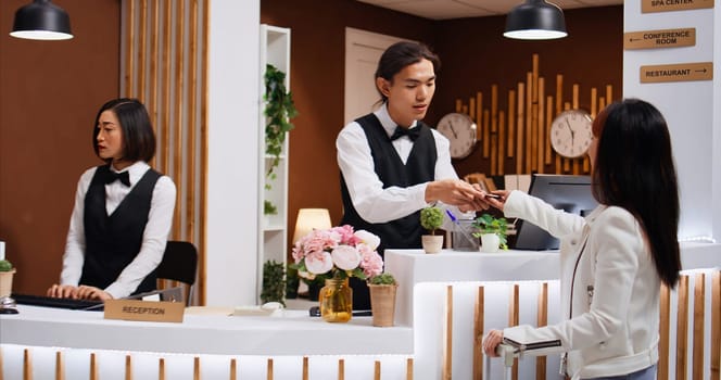 Hotel staff examines passport info for customer identification, finishing check in process at front desk. Receptionist verifying travelling id documents for asian woman arriving at reception.