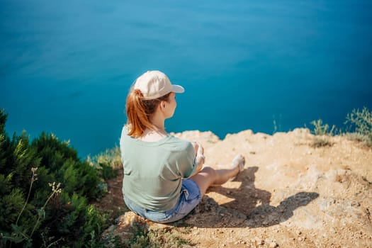 A woman is sitting on a rock overlooking the ocean. She is wearing a hat and a green shirt