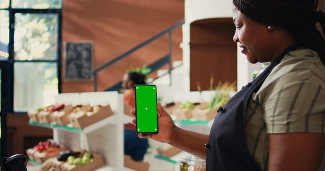 African american vendor holding phone with greenscreen, presenting isolated chromakey layout on device at farmers market checkout. Woman using copyspace screen on smartphone. Tripod shot.