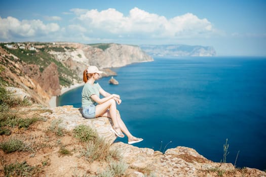 A woman sits on a rock overlooking the ocean. The sky is clear and the water is calm. The woman is wearing a green shirt and white hat