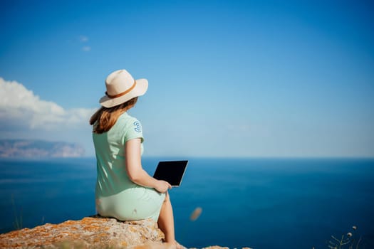 A woman is sitting on a rock by the ocean with a laptop in front of her. She is wearing a straw hat and a green dress. Concept of relaxation and leisure
