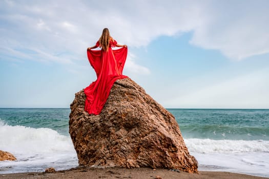 woman sea red dress. Woman with long hair on a sunny seashore in a red flowing dress, back view, silk fabric waving in the wind. Against the backdrop of the blue sky and mountains on the seashore