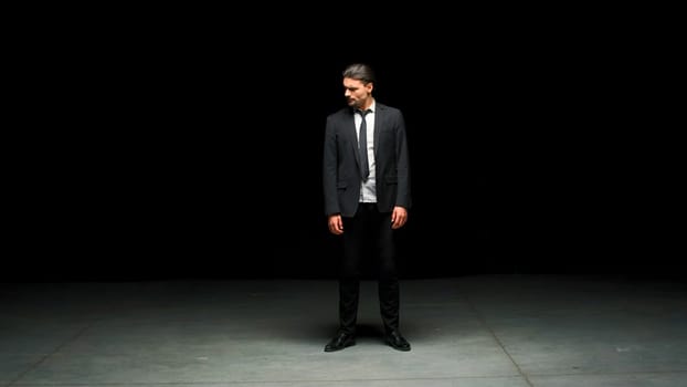 Man in suit stands emotionally on stage. Stock footage. Man in suit stands on stage with dramatic look. Elegant dramatic man on dark stage in emotional production.