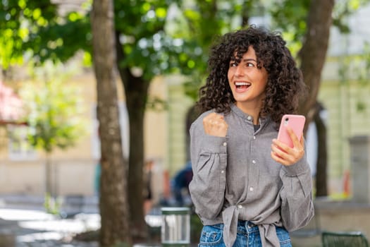 Caucasian young woman use mobile smartphone celebrating win good message news, lottery casino jackpot victory, giveaway online outdoors. Lady girl on urban city street. Town lifestyles. Sunny park