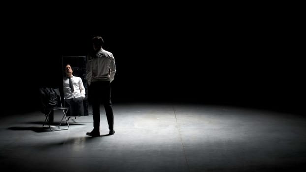 Man in suit in front of mirror. Stock footage. Man on stage with mirror puts on suit. Stage production with one man and mirror on dark stage.