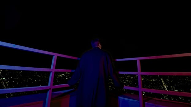 Stylish young man looks at city from roof. Media. Fashion man in coat on roof of skyscraper overlooking city. Young man looks at night city from roof of high-rise building.