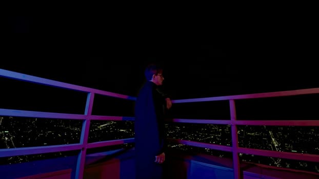 Stylish young man looks at city from roof. Media. Fashion man in coat on roof of skyscraper overlooking city. Young man looks at night city from roof of high-rise building.