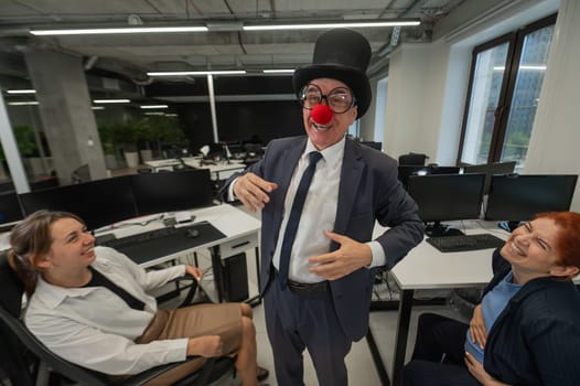 An elderly Caucasian man in a clown costume amuses two Caucasian women in the office