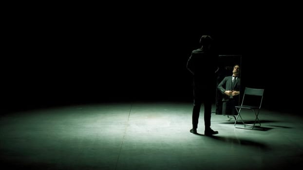 Man in suit in front of mirror. Stock footage. Man on stage with mirror puts on suit. Stage production with one man and mirror on dark stage.