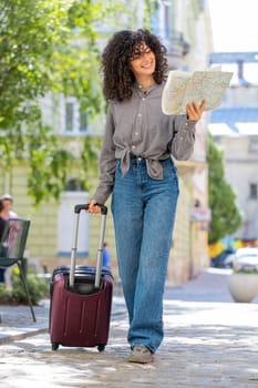 Young woman tourist with luggage suitcase exploring checking paper map search a way direction while traveling abroad. Lady girl walking on urban summer city street. Town lifestyles outdoors. Vertical