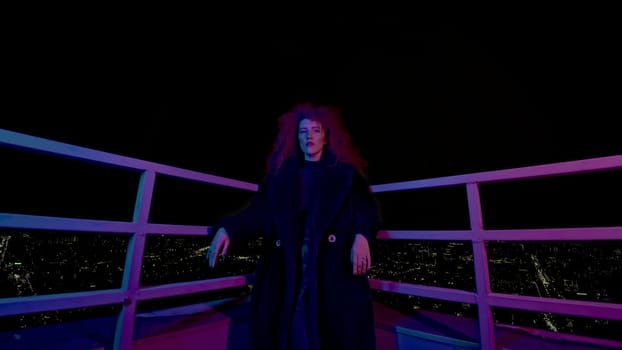 Woman on roof of night city. Media. Stylish fashion shoot with model in coat on roof. Female model in neon night shooting on roof of high-rise.