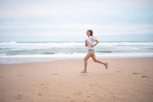 Full length of athletic woman running along seashre. Side view of young sportswoman in white top and sporty shorts jogging on sandy beach. Fit lady represents healthy lifestyle.