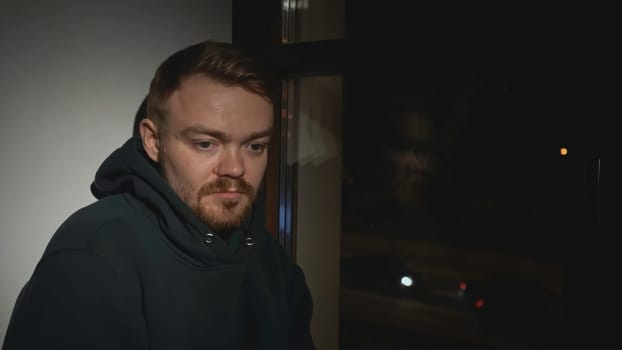 Sad man at night window of house. Media. Man looks out window with sad look and goes to sleep. Tired lonely man looks out window of house.