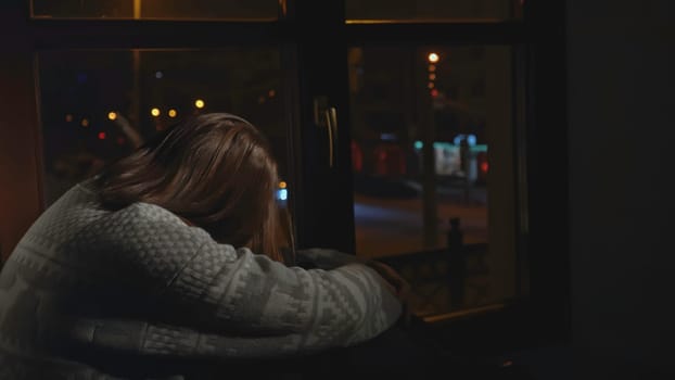 Lonely young woman looks out night window. Media. Young woman sits on window and looks sadly at night city. Woman with depression sits alone on window of house.