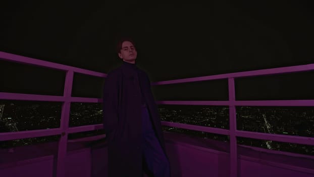 Young man stands on roof of building at night. Media. Handsome man in coat is standing on roof of high-rise building. Photo shoot of man on roof of high-rise building.