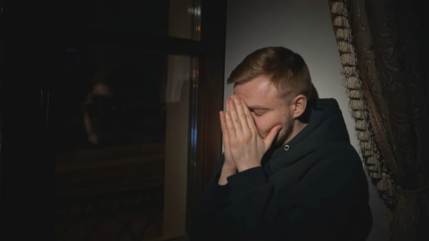 Young man sits tired and looks out window. Media. Lonely man looks out window of house and is sad. Man is nervous and sad looking out night window of house.