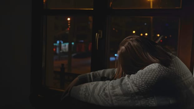 Lonely young woman looks out night window. Media. Young woman sits on window and looks sadly at night city. Woman with depression sits alone on window of house.