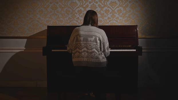 Woman starting to play piano. Media. Rear view of young woman playing piano. Woman in old sweater plays old piano.