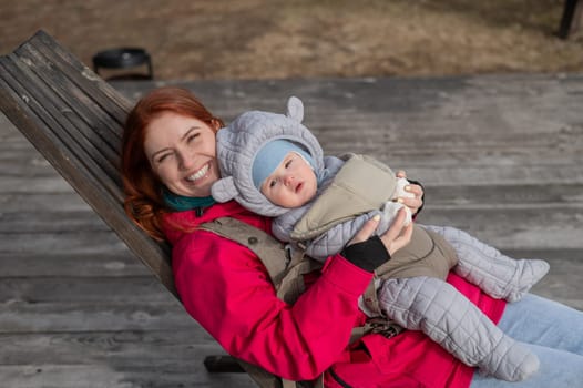 Caucasian woman with her son in an ergo backpack sitting in a wooden deck chair