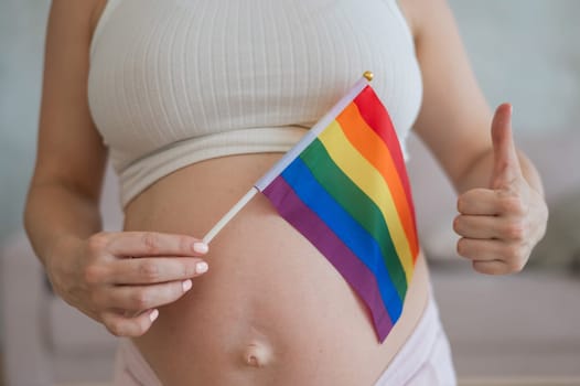 A woman holds a rainbow flag against the background of a naked pregnant belly and shows a thumbs up