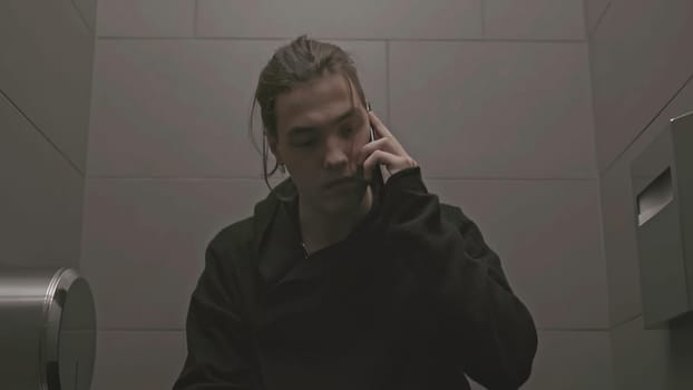 Young man sits on toilet and talks on phone. Stock. Man in black walks into toilet stall and talks on phone. Young man is talking aggressively on phone in toilet.