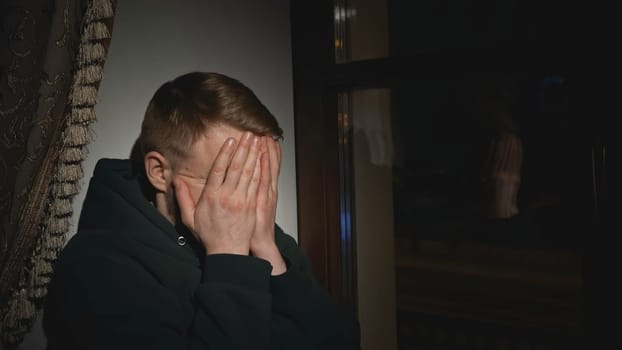 Young man sits tired and looks out window. Media. Lonely man looks out window of house and is sad. Man is nervous and sad looking out night window of house.