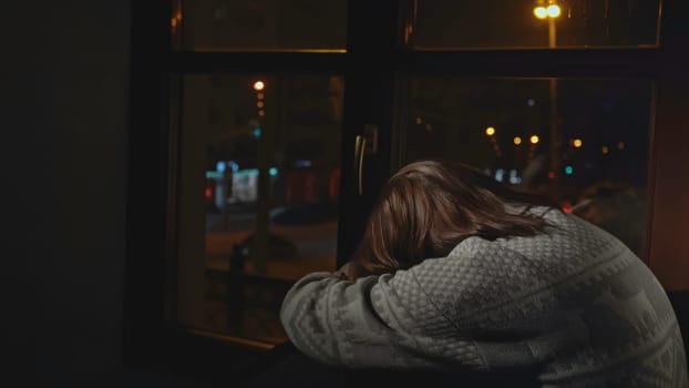 Lonely young woman looks out night window. Media. Young woman sits on window and looks sadly at night city. Woman with depression sits alone on window of house.