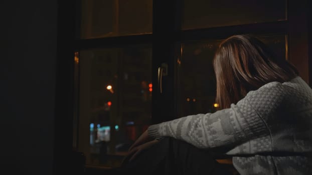 Rear view of woman sitting sadly by window. Media. Woman in sweater sits alone by window. Depressed young woman is sad on horse at home at night.