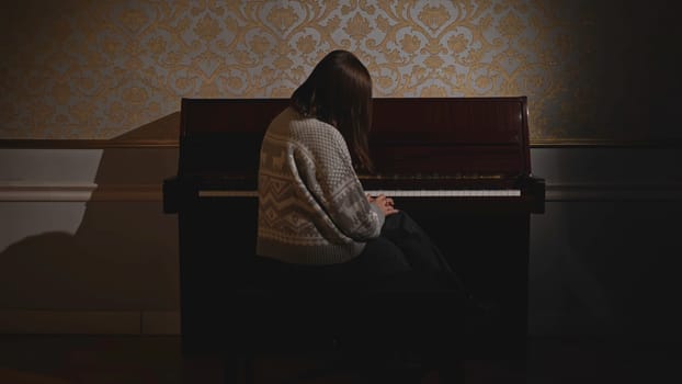 Young woman sitting sadly at piano. Media. Rear view of woman sitting at piano. Woman sits sadly at old piano and does not play.