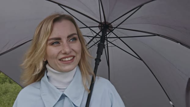 Young blond woman holding grey umbrella outdoors on green meadow. Stock clip. Rainy weather day using umbrella