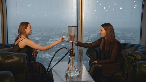 Young women with hookah and champagne in restaurant. Media. Two friends celebrate in restaurant with champagne and hookah. Attractive girlfriends relax in restaurant with hookah.