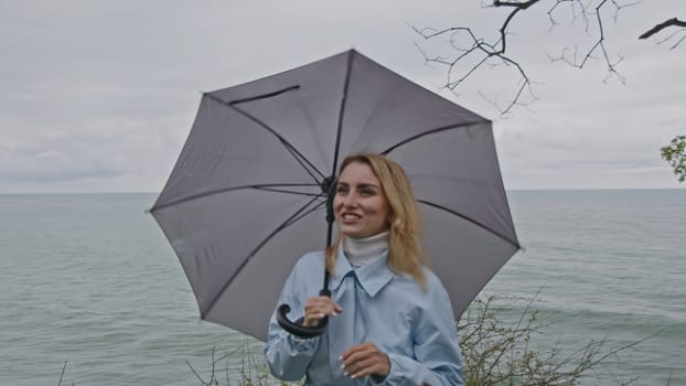 Girl in a light blue coat with a grey umbrella by the sea. Windy weather, green grass and rippling waves on the background