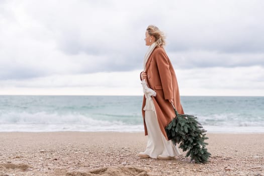 Blond woman Christmas sea. Christmas portrait of a happy woman walking along the beach and holding a Christmas tree in her hands. She is wearing a brown coat and a white suit