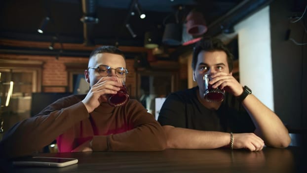 Young men are sitting at bar chatting and drinking cocktails. Media. Friends are sitting in cocktail bar and talking. Men relax at bar with cocktails.