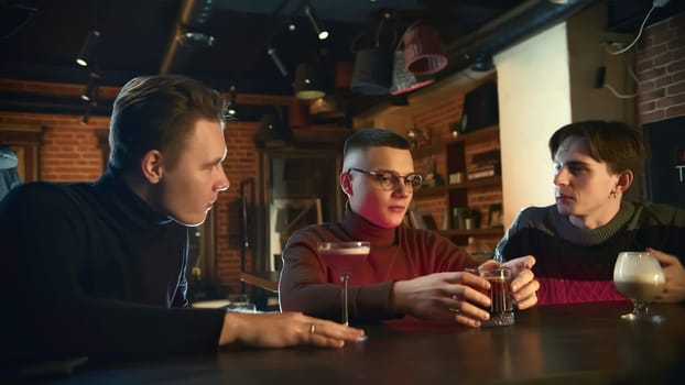 Young men are sitting at bar chatting and drinking cocktails. Media. Friends are sitting in cocktail bar and talking. Men relax at bar with cocktails.