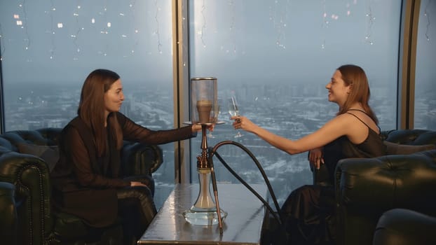 Young women with hookah and champagne in restaurant. Media. Two friends celebrate in restaurant with champagne and hookah. Attractive girlfriends relax in restaurant with hookah.