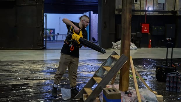 Young male worker using construction blower to clean assembled wooden stairs and pillar from sawdust, wood shavings. Media. Preparation of the stage for the concert