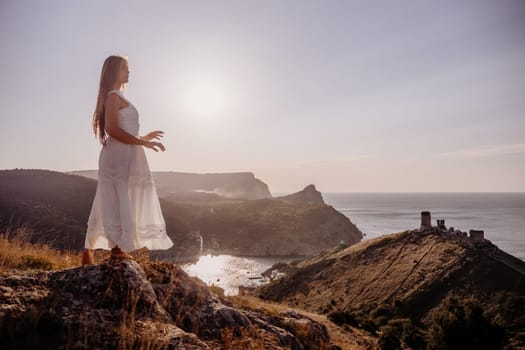 A woman stands on a rocky hill overlooking the ocean. She is wearing a white dress and she is enjoying the view. The scene is serene and peaceful, with the sun shining brightly in the background