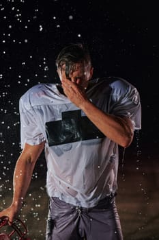 American Football Field: Lonely Athlete Warrior Standing on a Field Holds his Helmet and Ready to Play. Player Preparing to Run, Attack and Score Touchdown. Rainy Night with Dramatic Fog, Blue Light.