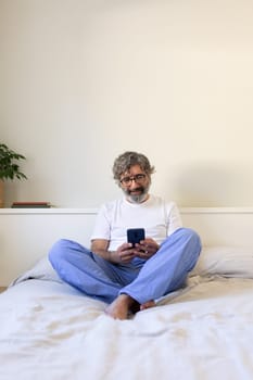 Vertical portrait of mature man using phone relaxing on bed at home. Copy space. Lifestyle concept.