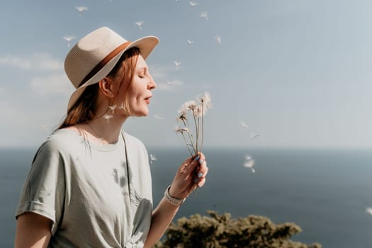 Young woman in a straw hat is standing on a beach and holding a bouquet of dandelion flowers. She is blowing the petals into the air, creating a beautiful and serene scene