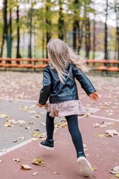 Little girl with flowing hair runs across the sports field towards the rows of seats. Back view. High quality photo