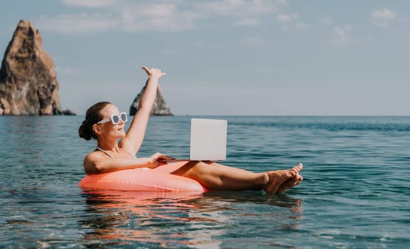 Woman freelancer works on laptop swimming in sea on pink inflatable ring. Pretty lady typing on computer while floating in the sea on inflatable donut at sunset. Freelance, remote work on vacation