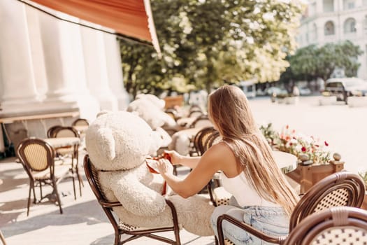 A woman sits cafe with a teddy bear next to her. The scene is set in a city with several chairs and tables around her. The woman is enjoying her time at the outdoor cafe