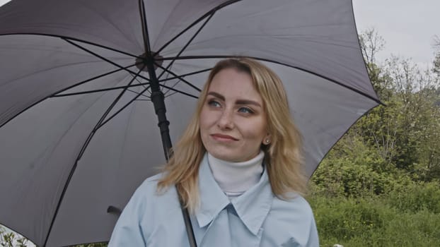 Portrait of cute romantic blonde woman smiling in green spring park. Stock clip. Young woman with grey umbrella
